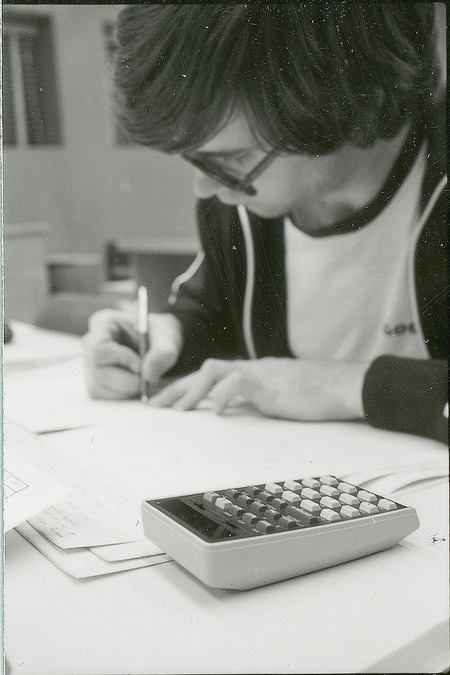 A young man is seated at the table on which there are several sheets of paper and a calculator. The man is writing information on one of the sheets of paper. This photograph was featured in the Chemical Department's descriptive brochure for 1978.