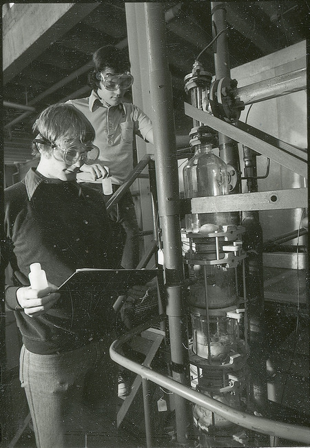 Two researchers in safety goggles are working with a piece of equipment. One researcher is carrying small plastic bottle and log book. This photograph was featured in the Chemical Department's descriptive brochure for 1978.