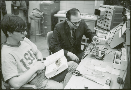 Two researchers work on the circuits of an apparatus. One researcher is consulting a large book, while the other is using a probe to test the circuits.