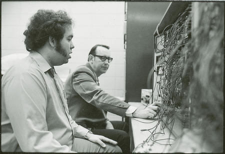Professor Burkhart and another researcher are seated before a control panel with square gauges. In the fore ground there is a mass of coated wiring.