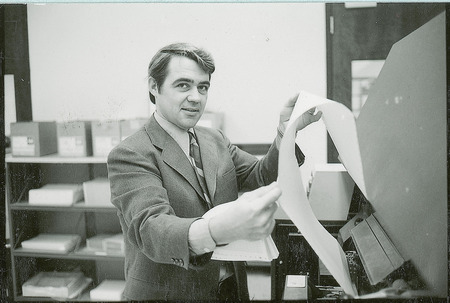 A researcher is holding a paper printout that is still in the process of being printed. Instead of looking at the printout however, he is looking directly at the photographer.