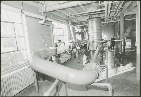 A researcher wearing both safety glasses and noise reduction muffs studies a section of tubing. This tubing is part of a larger piece of equipment.