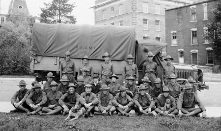 A unit of U.S. soldiers from Missouri, the "Motor Truck Second Section." Twenty seven men and two dogs are posed for a photograph by a military transport vehicle. The truck's canvas cover is stenciled with the insignia "U.S.Q.M.C." The tower of the Laboratory of Mechanics, the Student Services Building, and the West Boarding Cottage can be seen in the background.