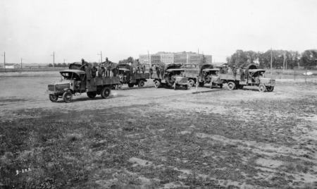 A U.S. army detachment in training. Five military transport trucks are parked on a field, each with canvas tops rolled up and five men in or on the truck. An officer is pointing at one of the trucks.