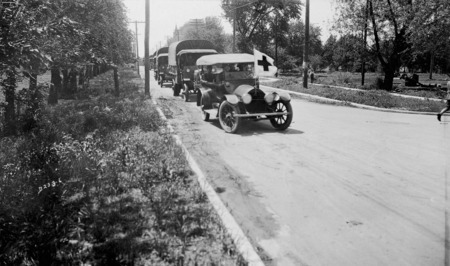 A U.S. army convey about to turn a corner on a city street. The convoy is led by a civilian vehicle with a red cross flag mounted on the front end.