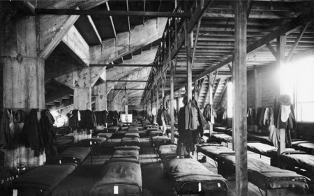A military training barracks. Rows of cots are positioned under a sports stadium structure. This is possibly the North Stadium.