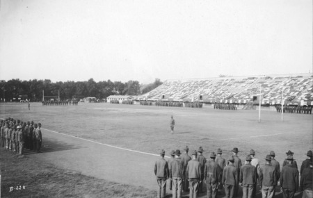 A military parade drill on the Iowa State football field. Platoons are standing at attention on the track surrounding the field with the exception of one platoon at the end of the field. Two officers stand at attention on the field. The West Stadium is in the background.