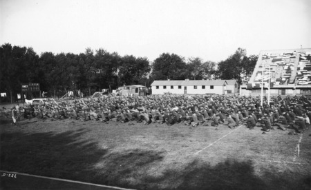 A large body of soldiers engaged in physical exercise, with the drill leader on a platform at the front of the formation. The majority of the soldiers are positioned in the south end zone of the ISU football field.One end of the West Stadium is in the background. A goal post and the score board are pictured.