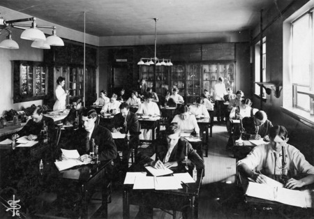 Students are writing at their desks in the Botany Lab in Central Building (Beardshear Hall). Some students have microscopes on their desks. Dr. Pammel, wearing a suit, is assisting a student at the back of the lab. A man wearing a white jacket is assisting another student. Estella Fogel (Buchanan) may be the woman standing at the left.