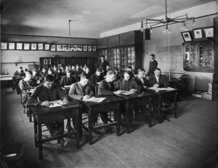 Dr. J.C. Cunningham, at the desk, is observing a class of all men in the Botany Lab in Central Building (Beardshear Hall). Mr. H.S. Coe, graduate assistant in the Botany Dept., is standing near the back of the room. Some men are sharing a desk and class materials.