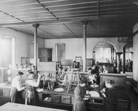 Men and women in a college chemistry laboratory. An adjoining laboratory, full of students, can be seen through a double arched doorway. A female student in the foreground appears to be wearing one of the men's hats.