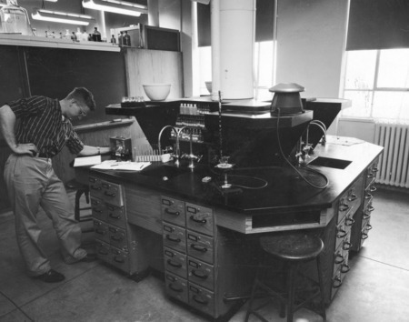 A student working in a newly designed laboratory for instruction in general chemistry employing semi-micro techniques. Test tubes and reagent bottles are arranged neatly at the work station. Four conjugate work stations are built as an island. This longer shot is very similar to the close up shot no. 1052-5-2.