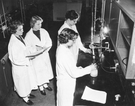 Four female students in white lab coats conducting an experiment in a chemistry laboratory. Two of the students are at the counter and two are to the rear watching while taking notes.