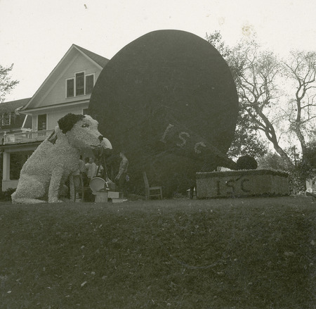 Cy the Dog sits attentively listening to a vintage phonograph player in this lawn display. A large dark disc is located behind the dog and phonograph. Several people are working on the display.
