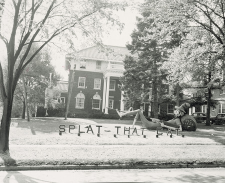 This lawn display consists of a semi-reclining bare-foot male figure dressed in ragged pants, shirt and hat. His head rests against a liquor barrel. He is pointing a shotgun at a Tiger figure that is suspended by one ear from a goal post.