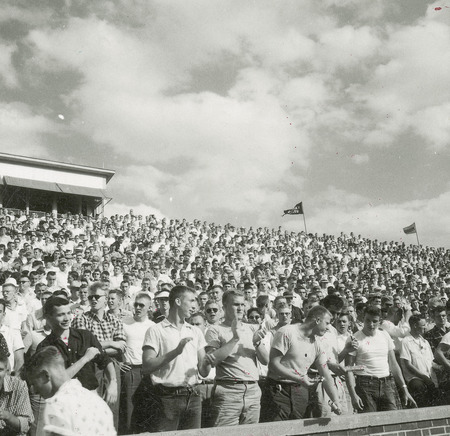 A group of spectators are standing in bleachers watching a Homecoming activity, quite possibly the football game.