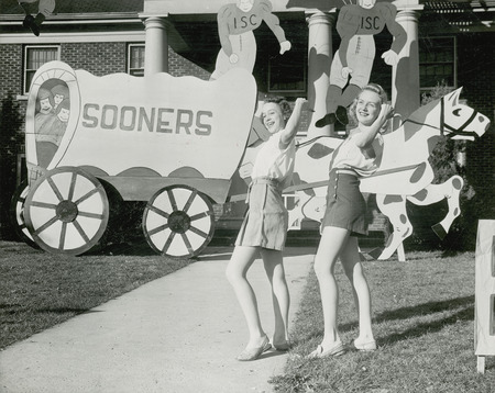 Two girls are posed in front of a lawn display depicting a horse-drawn covered wagon that has the words "Sooners" on the side. Two cardboard ISC football players are at the top of the photograph.