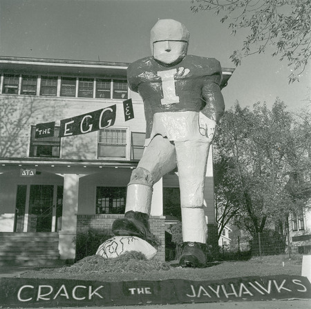 A sign reading "The egg &" points to the "I" on the jersey of a large football player standing on the lawn of the Delta Tau Delta, Gamma Pi Chapter house. The player is stepping on an egg and a banner reads "Crack the Jayhawks.".