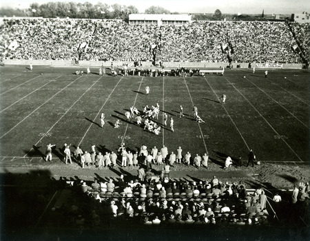 Football players on the field make a play during the Homecoming football game, viewed looking east from high in the stands, 1959.