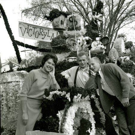 Homecoming attendant Joannie Parizek and two men lean on the Victoryland homecoming lawn display created by Phi Delta Theta, Iowa Gamma Chapter. The display won the Sweepstakes trophy, October 1961.
