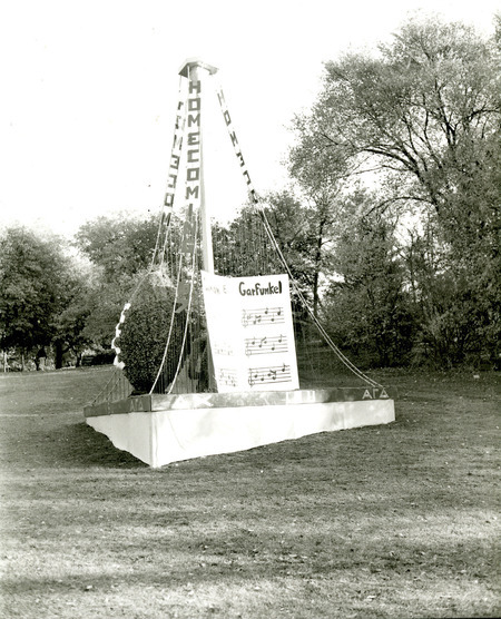 A Homecoming monument is shown that was built for the first time in 1967 by the Delta Chi and Alpha Gamma Delta. It has harp-like wires that drape over a football and a music score from Simon & Garfunkel.