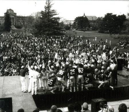 The Homecoming Pep Rally on Central Campus features the football team, viewed from behind the stage, 1977.