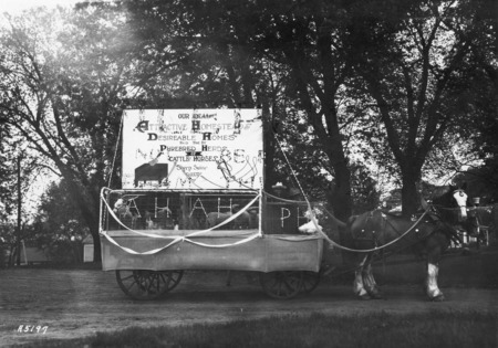A 1923 VEISHEA Animal Husbandry float. The horse-drawn float is mounted on a flat bed wagon that contains lambs, chickens, and a calf. The sign above reads: "Our Ideal Attractive Homesteads Are Desirable Homes Paid For By Purebred Herds of Cattle Horses Sheep Swine Poultry." See photograph 22-12-G.Veishea.1722-52 for alternate view.