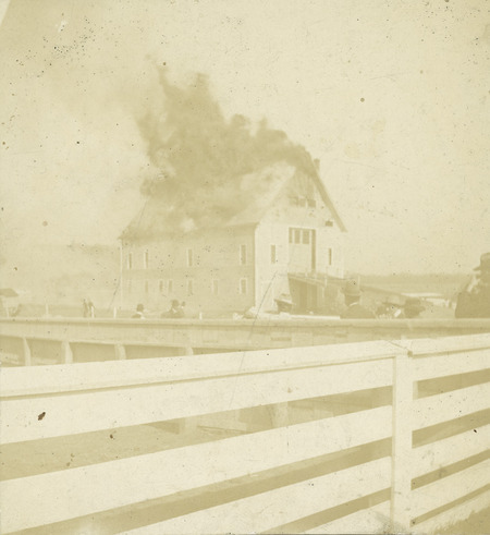The fire at the Iowa Agricultural College Experiment Barn in 1901. The building was of frame construction with a gable roof. Flames are engulfing the roof as a number of onlookers watch. 