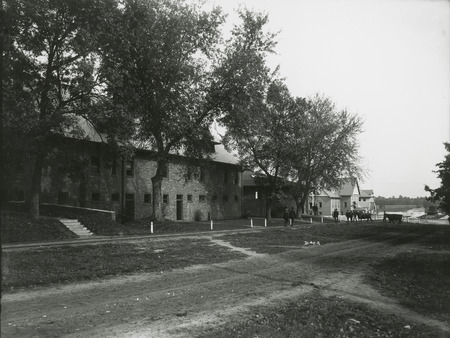 The brick Horse Barn on the left, built around 1900, with the Iowa Agricultural College Experiment Barn and the Judging Pavilion on the right. Two beef cattle are shown being led into the Pavilion as several men also approacch the area. 