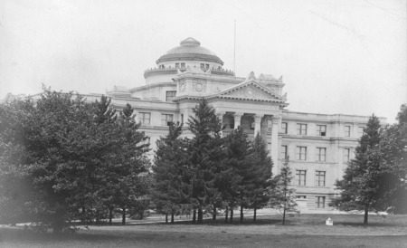 The front of Beardshear Hall as observed through the early growth tree groupings on central campus. An apparent weather station box sits on the lawn in front of the building.