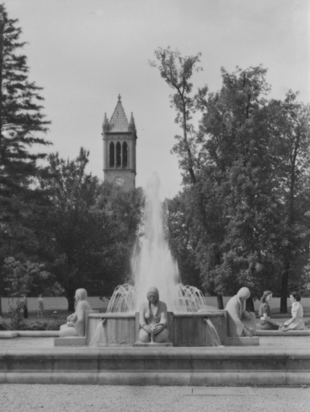 The Memorial Union fountain (Christian Petersen's Fountain of the Four Seasons) in spring with the Campanile towering over the trees in the background, the clock shows 2:50. Two young women are seated on the fountain edge on the right of the image.