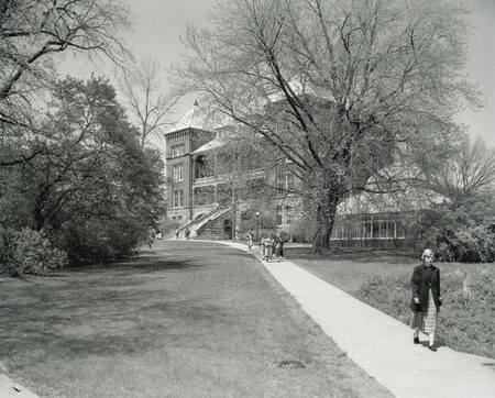 The front and side of Catt Hall. The greenhouse can also be seen through the trees. In the foreground, individuals can be seen walking along the sidewalk.