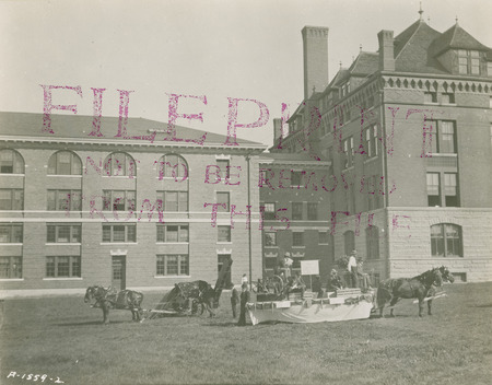 Several men on a float hitched to a pair of horses. Two other horses are hitched to a piece of farming equipment. The west-side of the original building and the 1903 north addition are in the background. The photograph has been stamped in ink "FILEPRINT NOT TO BE REMOVED FROM THIS FILE."
