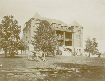 The front of Catt Hall although part of the building is obscured by trees. An artillery cannon and railroad tracks are in the foreground. Several individuals in period dress are standing on the stairs and sidewalk in front of the building.