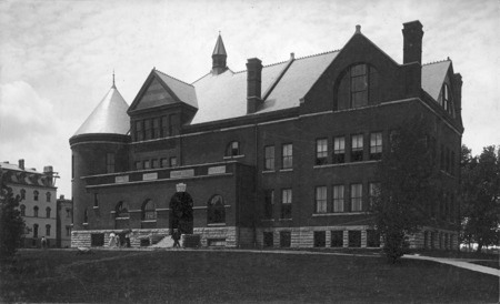 View of the front (east) and north sides of Morrill Hall. Three woman, one with an umbrella, are walking northward and two men are walking southward across the front of the building. Old Main is on the left side of the photograph. 