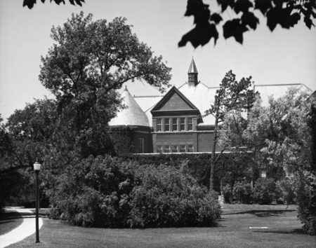 View of the front (east) side of Morrill Hall. Most of the view of the building is hidden by bushes and trees, so that the middle and upper parts of the building are most of what can be seen.