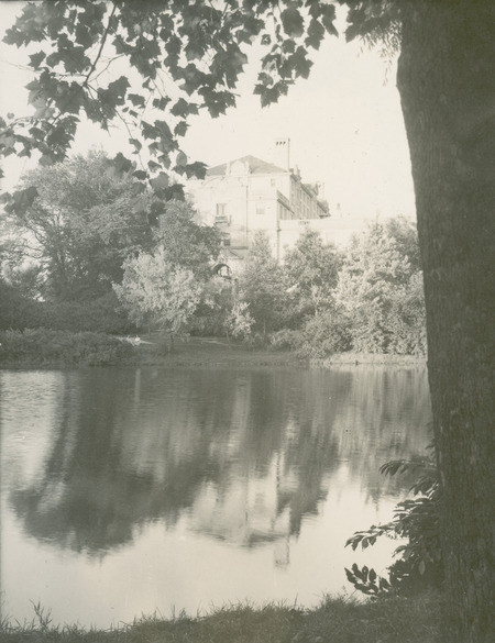 This 1944 photograph of the Memorial Union's west side shows mainly the roof lines, chimneys, pediments, a balcony around a window above the entrance, and the windows on the upper floors. Lake LaVerne is in the foreground, and plantings and trees hide most of the Union building. Two women are sitting on the far bank of the lake.