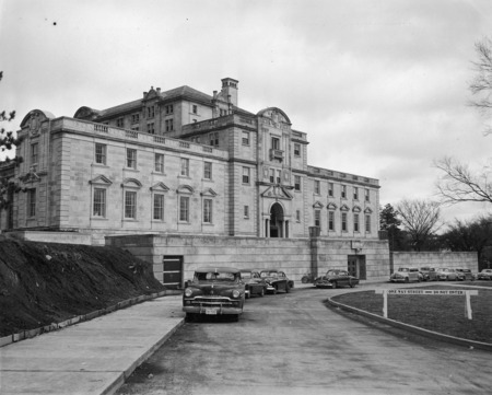 This 1953 photograph shows the entire west side and terrace of the Memorial Union from street level. In it are easily seen many of the Union's unique architectural details including the pediments over the windows and in the roofline, balustrades, chimneys and balconies. Several cars and a bicycle are parked along the curved street. The hill to the left is bare and un-landscaped, and a temporary barrier runs along its foot to keep dirt from spilling onto the sidewalk.