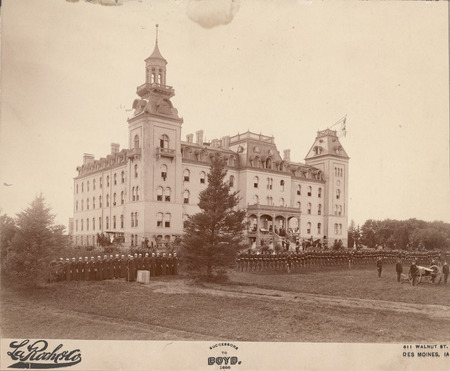 the front side of Old Main, with the college military cadet corps arranged in a review formation on the lawn in front of the building. An officer on a horse is at the foot of the Old Main steps. Military training was compulsory for male Iowa State students from 1868 through 1962.