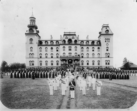 The front side of Old Main, with the college military cadet corps arranged in a review formation on the lawn in front of the building. The women's military corps is in front of the corps, in addition to a band and a drum major. Military training was required of Iowa State male students from 1868 through 1962 and women also organized a military corps from 1878 through 1897.