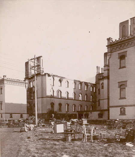 The north side of Old Main after a fire in 1900. A large number of chairs are strewn around the grounds. This view shows clearly that the north roof and north wall of the building are gone. Window openings on the west and south side of the north wing are empty of glass and framing.