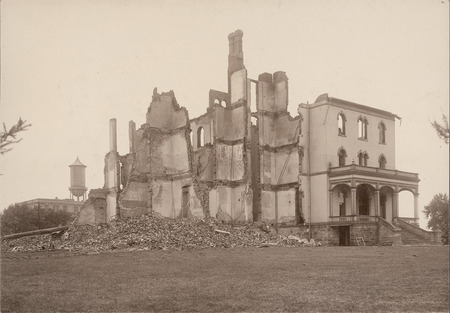 Old Main from the southeast after the second fire in August 1902. The gutted building stands behind a large heap of rubble. Only the front porch is still intact. The water tower is seen to the left of the building in the middle distance.