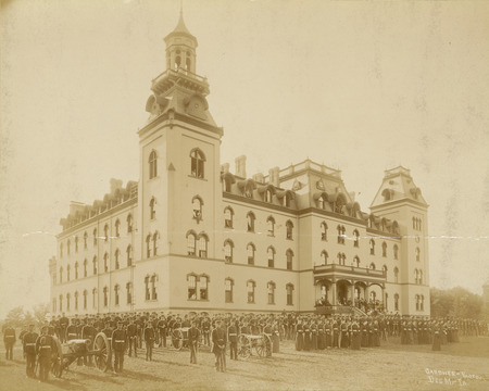 Old Main as the setting for a military corps review. Men's and women's corps are at attention. Military service for male students was mandatory between 1868 through 1962. For women, training was offered from 1878 through 1897. Three cannon are in the formation. An audience is on the stairs and front porch of Old Main.