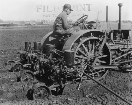 A four-row corn planter being pulled by a steel-wheeled tractor. A driver is at the wheel of the tractor. Buildings can be seen across a road in the distance. The photograph was taken April 27, 1931.