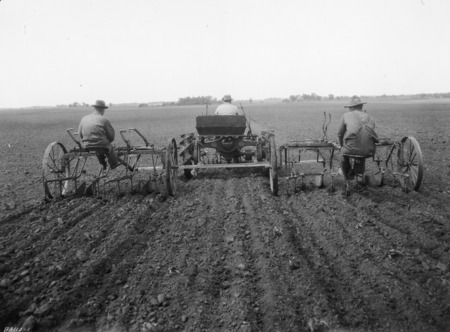 A tractor-mounted six-row cultivator seen from the rear. One man is driving the tractor.One man is also seated on each of the two rear sections of the cultivator, one on each side of the machine. The machinery and the tractor have steel wheels. They are in the middle of a very large plowed field.