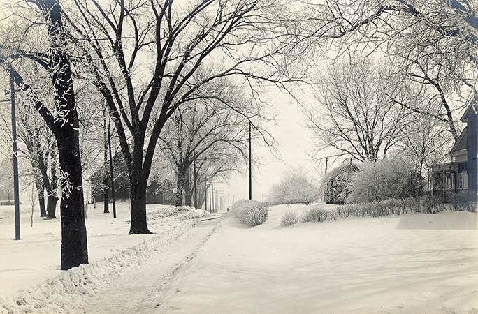 Photograph of a pathway cleared through the snow running east between the Farm House and Creamery, First (when it was probably known as the Farm Foreman’s Cottage) on the right (south side), and Landscape Architecture (when it was known as the Horse Barn) on the left (north side). Annotation: “Looking east Farm House on right, Horse Barn on left, after 1907 where the electric line turned north of Ag Hall & Margaret Hall.”