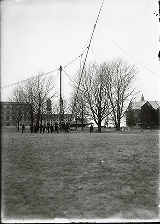 Photograph showing the raising of the flagpole on central campus. In the background Beardshear Hall, Marston Hall, the Marston Water Tower, the Hub, and Morrill Hall can be seen. Annotation: “Erecting of steel flagpole, ca. March 1908. Beardshear Hall, Marston Hall, water tower, Hub, and Morrill Hall in background.”