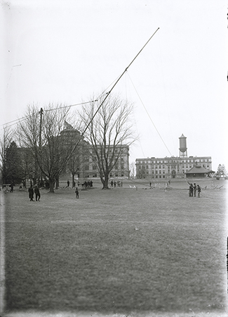 Photograph showing the raising of the flagpole on central campus. In the background Beardshear Hall, Marston Hall, the Marston Water Tower, and the Hub can be seen. Annotation: “Raising of flagpole. Bearshear Hall, Marston Hall, water tower & Hub, 1908.”