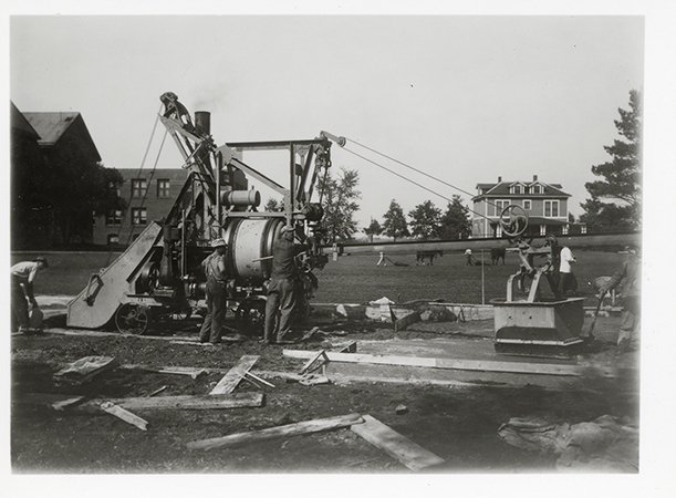 Photograph of people pouring cement for a driveway. The buildings in the background, from left to right (west to north) are Margaret Hall, North Hall, MacKay Hall, and the Bevier House. Annotation: “Flagpole, n.d.”