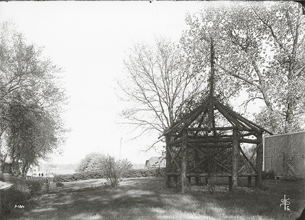 Photograph of an arbor with a barn behind it in the background. Annotation: “Summer House at Farm House. May 27, 1907.”